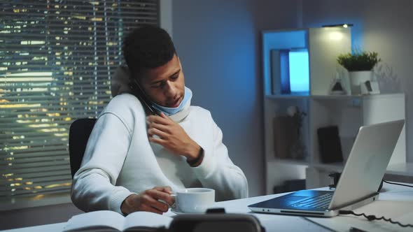 Medium Shot of Mixedrace Man Working on Computer Speaking By Smartphone and Drinking a Cup of Tea