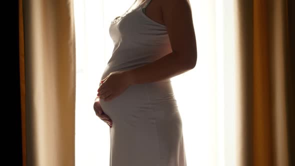 CLoseup of Pregnant Woman Stroking Her Growing Belly Against Big Window and Bright Sun Light