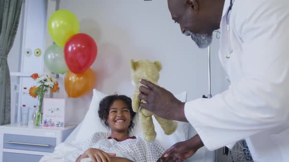 Diverse male doctor playing teddy bear with girl in hospital bed wearing fingertip pulse oximeter