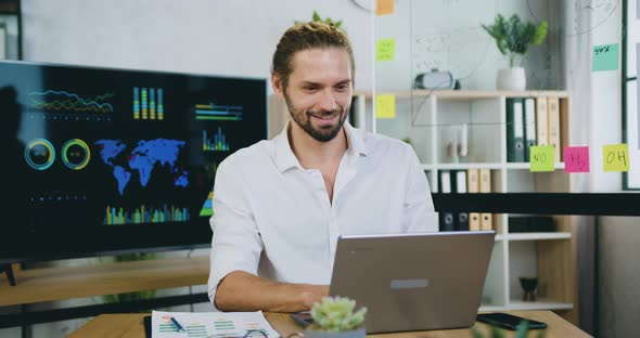 Young Employer Working at Laptop Sitting at a Table in front of a Computer Monitor