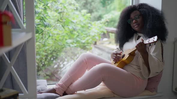 Wide Shot Talented Relaxed African American Woman Playing Ukulele Sitting on Windowsill at Home