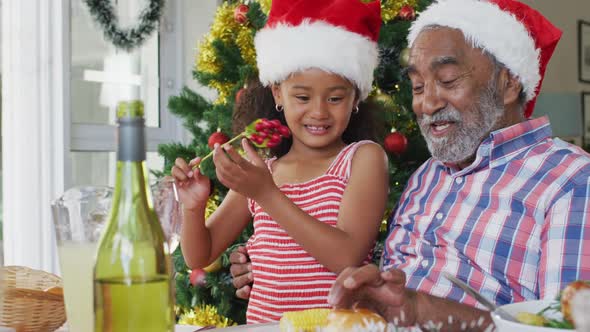 Happy african american grandfather and granddaughter wearing santa hats and celebrating at table