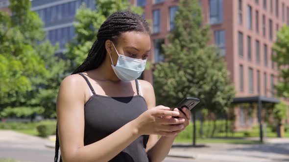 A Young Black Woman in a Face Mask Looks at a Smartphone and Celebrates - an Office Building