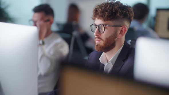 Serious Young Bearded Man Working on Decktop Computer While Working in Big Open Space Office