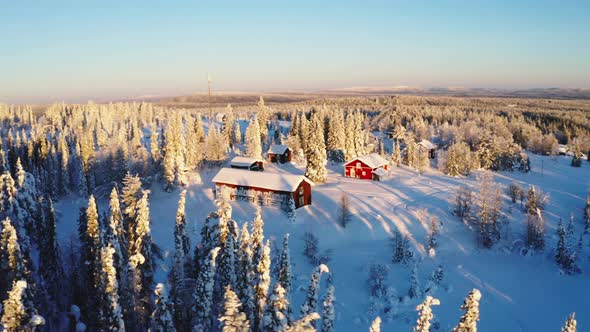 Aerial view across sunrise shining on snow covered red winter cabins surrounded by frosty woodland t