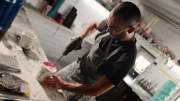 A male handyman mixes paint in a can in a workshop. A sculptor mixes resin with paint to a vase.