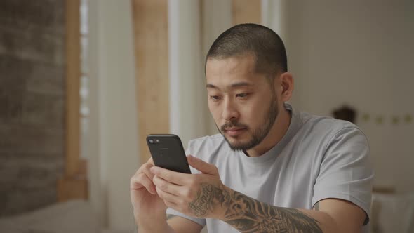 20s Tattoed Asian Man Browsing on His Smart Phone Indoors