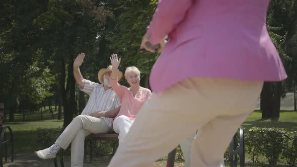Two Senior Men and One Woman Waving Hands While Their Friend Woman Driving on Scooter in the Park