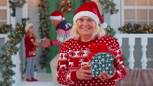 Portrait of Grandmother Woman Presenting Gift Box Smiling Near Decorated Christmas House with Family