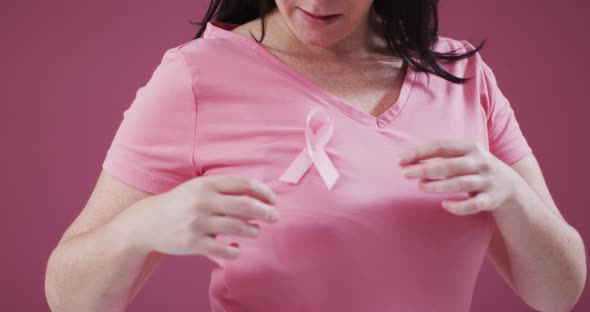 Mid section of a woman touching the pink ribbon on her chest against pink background