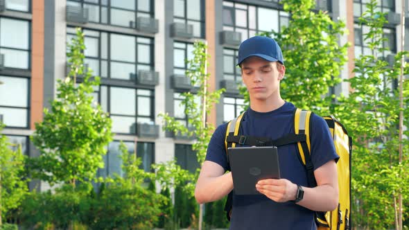 Portrait Young Man Courier Food Delivery with Thermal Backpack Uses Tablet