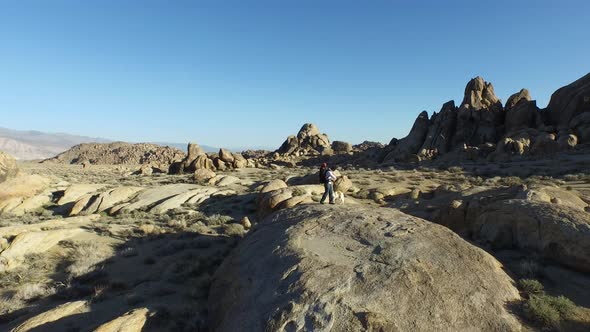 Aerial shot of a young man backpacker standing on a boulder with his dog in a desert mountain range.