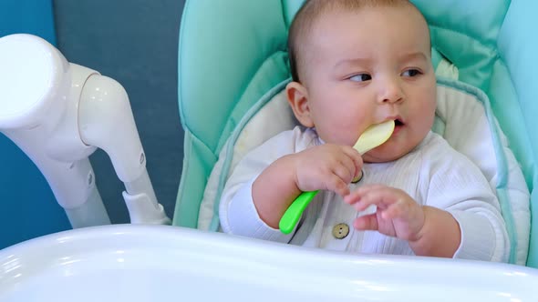 A hungry baby is gnawing on a plastic spoon at the table on a high chair. Teething, whims, itchy gum