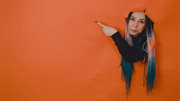 Young Woman Sticking Out of Hole of Orange Background and Pointing Index Finger at Space for Text