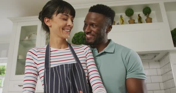 Happy diverse couple wearing blue apron embracing and cooking in kitchen