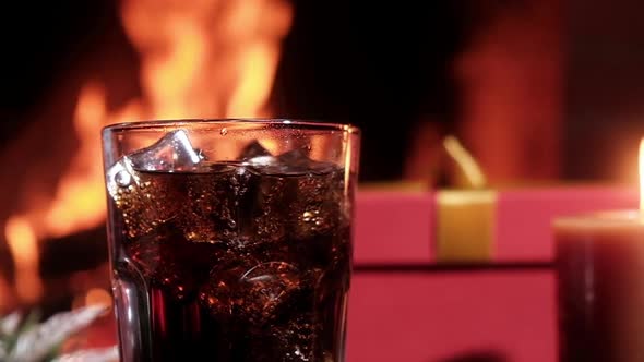 Carbonated Cola drink in glass with ice in front of burning fire in fireplace.