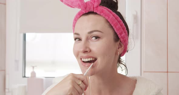 Beautiful Woman Using Oral Irrigator To Clean Her Teeth In A Bathroom Looking At Camera With Window