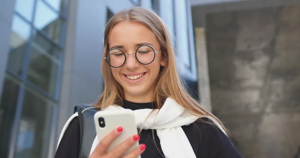 Young Trendy Blondie in Glasses which Standing Near Modern City Building and Using Her Smartphone