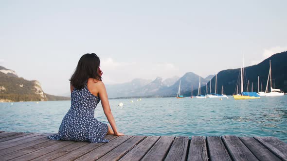 Young Woman in Dress Sitting at the Edge of Wooden Pier at Lake in Mountains Enjoying View