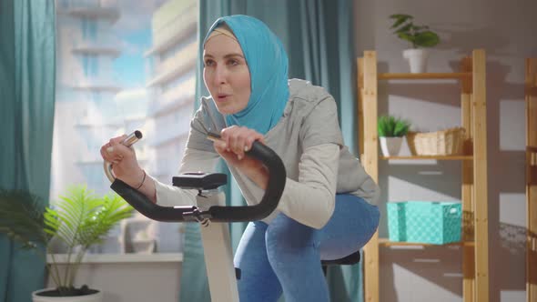 Young Muslim Athlete in a National Headscarf is Engaged on an Exercise Bike at Home