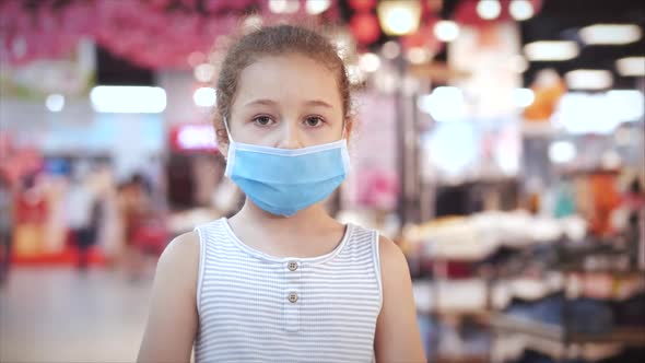 Cute Little Girl From an Epidemic of Coronaviruses or Viruses Looks at the Camera Amid Masked People