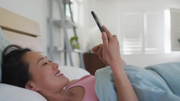 Happy asian woman lying in bed, using smartphone in the morning