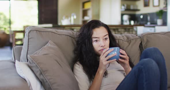 Relaxed biracial woman with vitiligo drinking coffee, sitting on sofa