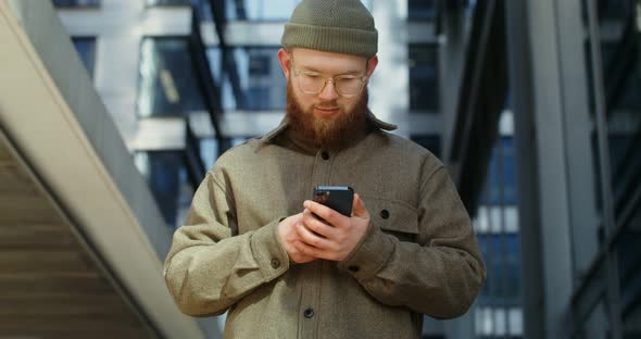 A Bearded Man is Typing on a Mobile Phone While Standing Near Office Building