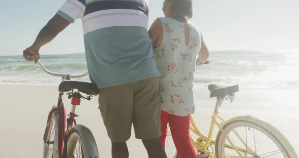 Senior african american couple walking with bicycles on sunny beach