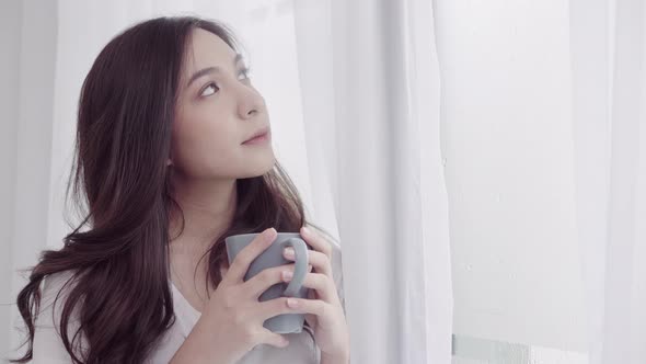 Asian woman smiling and drinking a cup of coffee or tea near the window in the bedroom.