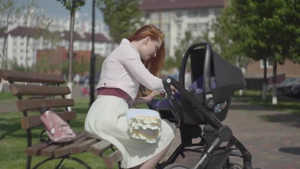 Portrait Red-haired Woman Playing with Her Kid Lying in the Pram in the Park Close-up. Mom Enjoying