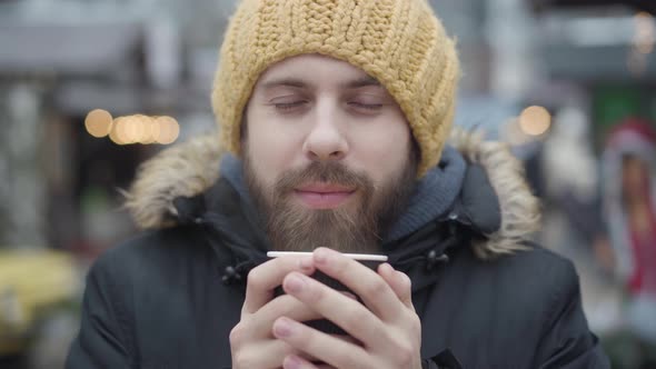Close-up Portrait of Frozen Young Caucasian Man Drinking Hot Coffee on the Street, Cheerful Guy 