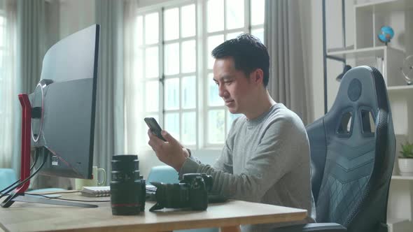 Asian Cameraman Using Mobile Phone While Using Desktop Computer For Working At Home