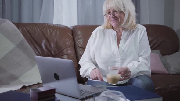 Positive Caucasian Retiree Sitting on Sofa with Coffee Cup and Looking at Laptop Screen