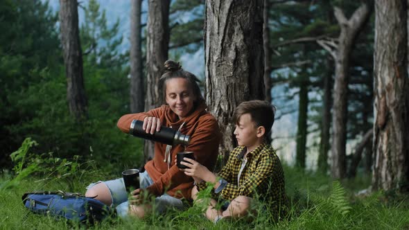 Mother with son relaxing in the forest and drinking hot herbal tea from a thermos on a sunny day