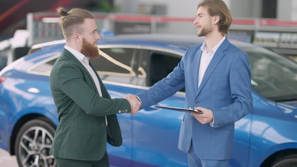 Side View of Millennial Caucasian Client and Dealer Shaking Hands and Passing Keys in Car Dealership