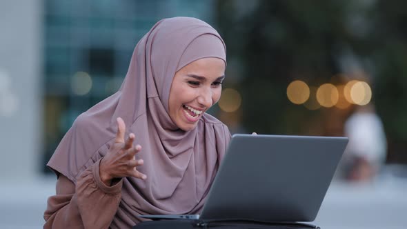 Excited Young Muslim Woman Winner Looks at Laptop Celebrates Online Success Sits Outdoors in City