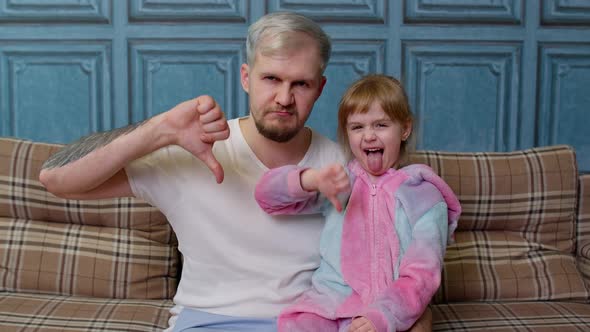 Displeased Father and Child Daughter Kid in Pajamas Sit on Couch in Room Showing Thumb Down Dislike