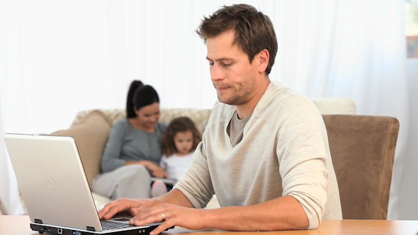 Man Working On His Laptop While His Family Is On The Sofa