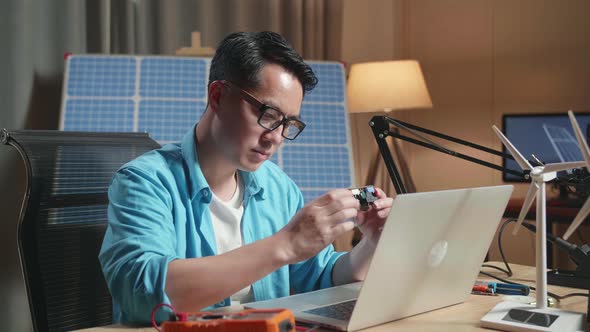 Asian Man With Wind Turbine Looking At Circuit Board While Working With Laptop