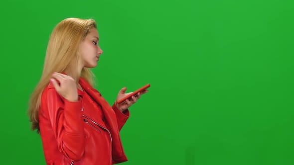 Portrait Blonde Girl Is Walking and Reading Text Message on Mobile Phone at Green Screen. Slow