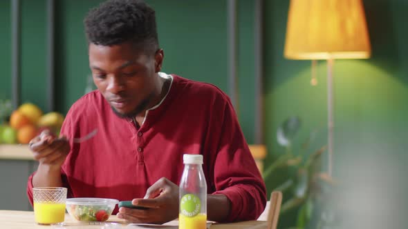 Young Black Man Having Lunch and Using Smartphone at Kitchen Table