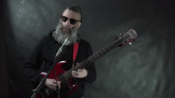 Bearded hairy rockstar plays on red electric guitar on stage with black background