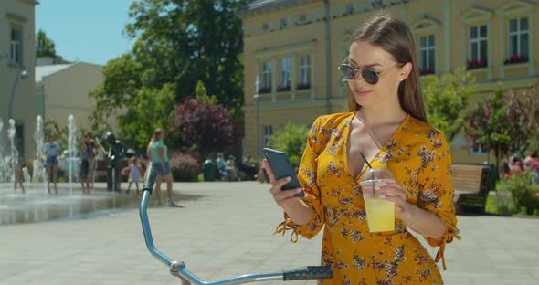 Young Woman on a Bicycle Drinking Lemonade and Using Smart Phone on the Square in Front of the