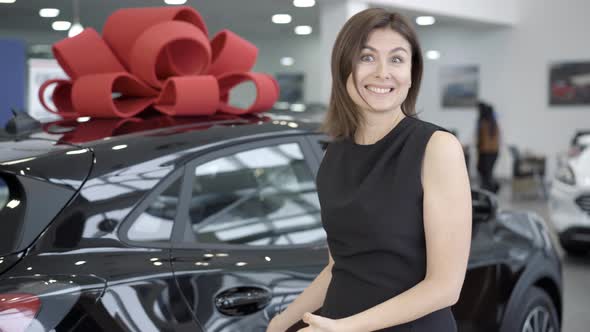 Portrait of Happy Young Woman Standing in Car Dealership and Cheering Purchase of New Black