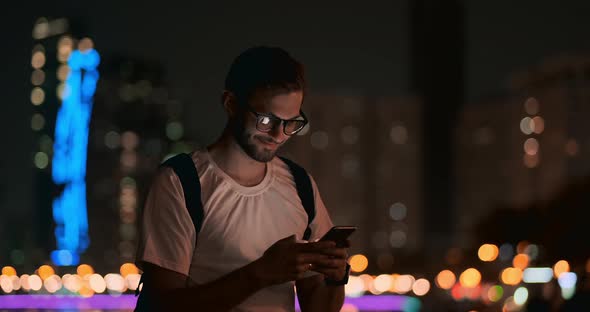 A Man in Glasses Against the Background of the Night City Writes a Message Using a Smartphone