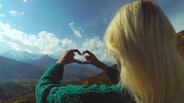 Woman on hike on top of mountain makes heart shaped finger frame with her hands.