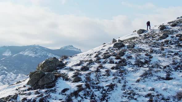 A Traveler with a Backpack Climbs the Slope of a Hill Covered with Snow Against the Backdrop of a