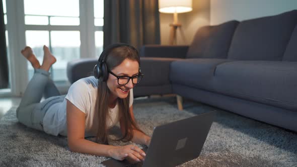 Woman Is Lying on the Floor, Working on a Laptop and Listens To Music on Headphones