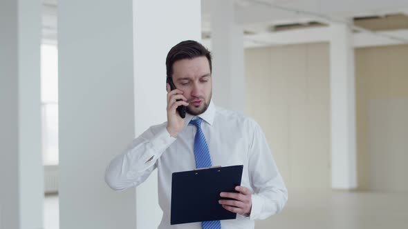 Realtor man with tablet in hands is standing in middle of an empty office space and talking on smart